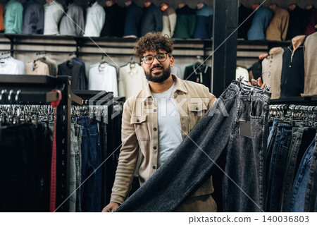 Male customer in a clothing store holds up a pair of dark jeans while browsing through a rack of apparel, with various shirts displayed in the background Male customer in a clothing store holds up a pair of dark jeans while browsing through a rack of apparel, with various shirts displayed in the background 140036803