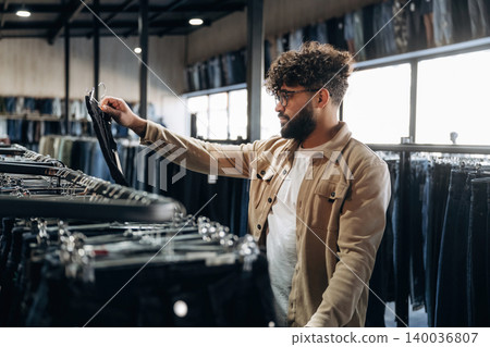 Male shopper with curly hair examines black jeans on a hanger inside a clothing store, racks of denim and bright windows visible in the background Male shopper with curly hair examines black jeans on a hanger inside a clothing store, racks of denim and bright windows visible in the background 140036807