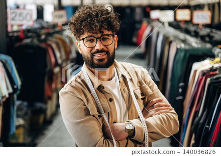 Male tailor with curly hair and glasses stands confidently in a clothing store, surrounded by racks of various garments and price tags visible in the background Male tailor with curly hair and glasses stands confidently in a clothing store, surrounded by racks of various garments and price tags visible in the background 140036826