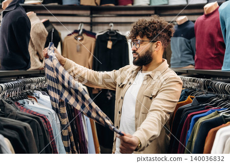 Male shopper examining a checkered shirt while browsing clothing racks filled with various garments in a retail store setting 140036832