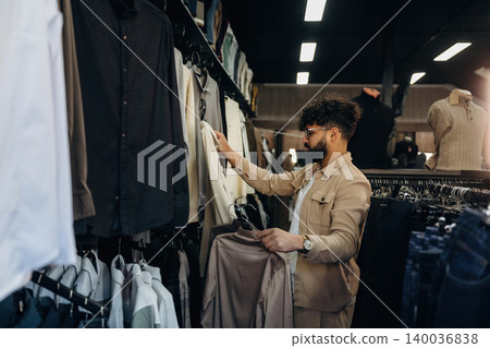 Male shopper with curly hair browsing clothing racks in a modern retail store, holding a beige shirt while examining various garments on hangers 140036838