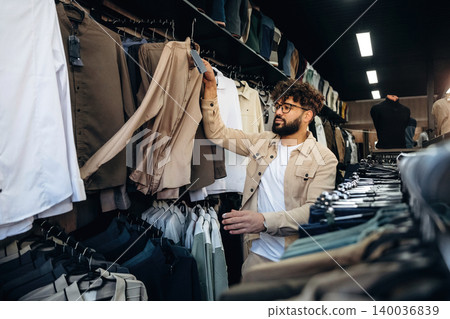 Male customer browsing clothing racks in a retail store, examining beige and light-colored garments, with various shirts and pants displayed on hangers 140036839