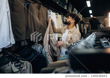 Male shopper with curly hair examines clothing in a retail store, surrounded by neatly arranged shirts and blouses on hangers in a modern fashion environment Male shopper with curly hair examines clothing in a retail store, surrounded by neatly arranged shirts and blouses on hangers in a modern fashion environment 140036841