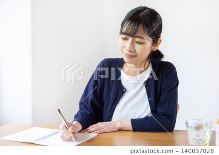A young Japanese woman is taking notes in a notebook at a table with a bottle of pills and a glass. A young Japanese woman is taking notes in a notebook at a table with a bottle of pills and a glass. 140037028