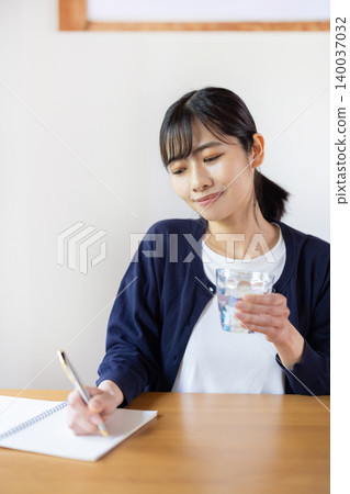 A young Japanese woman is taking notes in a notebook at a table with a bottle of pills and a glass. 140037032