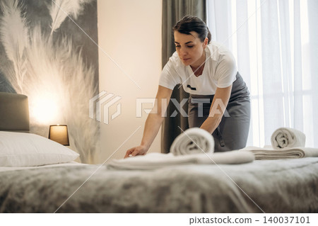 Hotel housekeeping staff member arranging towels on a neatly made bed in a modern guest room with soft lighting and elegant decor elements Hotel housekeeping staff member arranging towels on a neatly made bed in a modern guest room with soft lighting and elegant decor elements 140037101