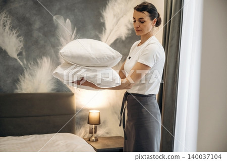 Female hotel staff member arranging white pillows on a neatly made bed in a modern guest room with decorative wall art and bedside lamp Female hotel staff member arranging white pillows on a neatly made bed in a modern guest room with decorative wall art and bedside lamp 140037104