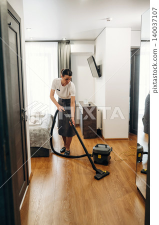Female hotel staff member vacuuming wooden floor in a modern hotel room with a bed, mirror, and television visible in the background Female hotel staff member vacuuming wooden floor in a modern hotel room with a bed, mirror, and television visible in the background 140037107