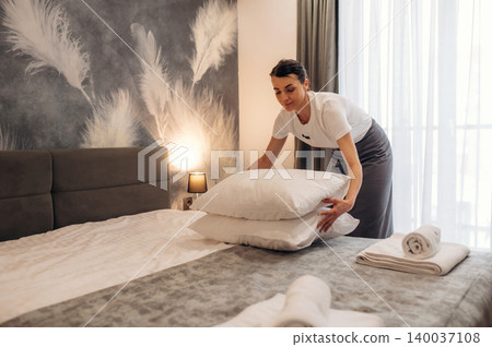 Hotel housekeeping staff member arranging pillows on a neatly made bed in a modern guest room with decorative wall and natural light from large window Hotel housekeeping staff member arranging pillows on a neatly made bed in a modern guest room with decorative wall and natural light from large window 140037108