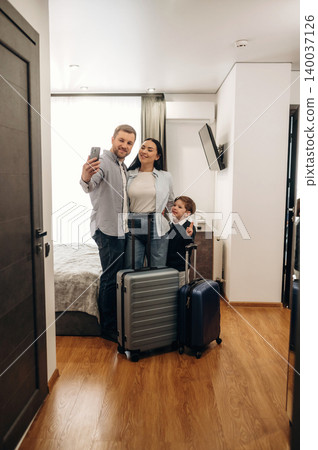 Family of three taking a selfie in a hotel room with luggage, featuring a double bed, wooden floor, and modern decor elements in the background Family of three taking a selfie in a hotel room with luggage, featuring a double bed, wooden floor, and modern decor elements in the background 140037126