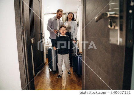 Family with luggage entering hotel room, young boy leading with suitcase, parents following, modern interior design visible in the background Family with luggage entering hotel room, young boy leading with suitcase, parents following, modern interior design visible in the background 140037131