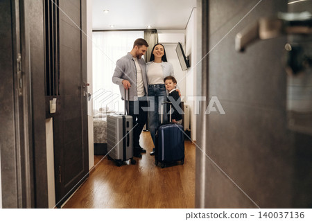 Family with luggage standing in hotel room doorway, modern decor visible, bright natural light streaming through large window, welcoming atmosphere created by warm wooden flooring Family with luggage standing in hotel room doorway, modern decor visible, bright natural light streaming through large window, welcoming atmosphere created by warm wooden flooring 140037136