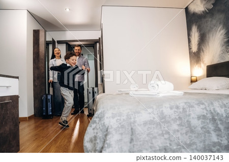 Family entering a modern hotel room with luggage, a child excitedly running towards the bed, and neatly arranged towels on the gray bedspread Family entering a modern hotel room with luggage, a child excitedly running towards the bed, and neatly arranged towels on the gray bedspread 140037143