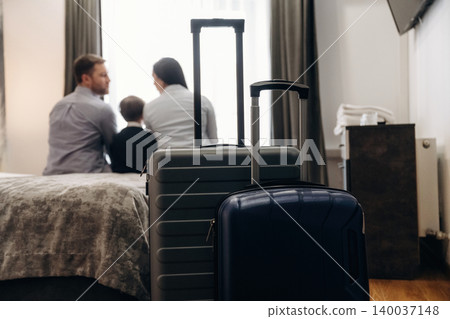 Family of three with luggage sitting on bed in hotel room, large window with curtains and television visible in the background Family of three with luggage sitting on bed in hotel room, large window with curtains and television visible in the background 140037148
