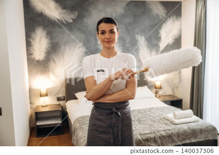 Female hotel housekeeper holding a duster in a modern hotel room with a bed, decorative pillows, and a stylish feather-patterned wall behind her Female hotel housekeeper holding a duster in a modern hotel room with a bed, decorative pillows, and a stylish feather-patterned wall behind her 140037306