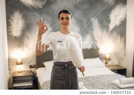 Hotel housekeeping staff member in a white shirt and gray apron holds a feather duster while standing in a well-decorated hotel room with a bed and lamps Hotel housekeeping staff member in a white shirt and gray apron holds a feather duster while standing in a well-decorated hotel room with a bed and lamps 140037310