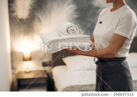 Female hotel staff member holding neatly stacked white towels and a rolled towel in a modern hotel room with a bed and bedside lamp visible in the background Female hotel staff member holding neatly stacked white towels and a rolled towel in a modern hotel room with a bed and bedside lamp visible in the background 140037315