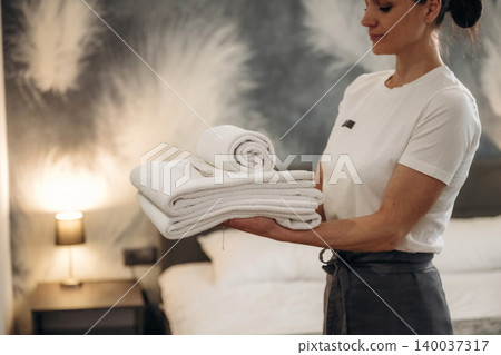 Female hotel staff member holding neatly stacked white towels in a modern hotel room with a stylish wall design and a bed in the background 140037317
