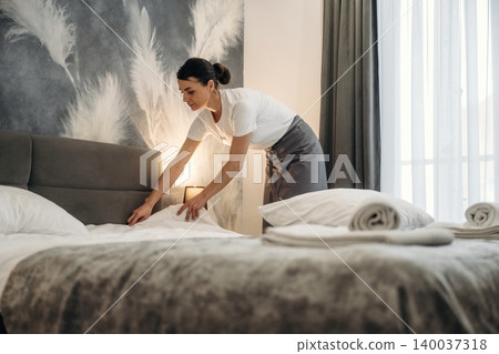 Hotel housekeeper arranging bed linens in a modern guest room with decorative wall and large window, showcasing a clean and inviting atmosphere for guests Hotel housekeeper arranging bed linens in a modern guest room with decorative wall and large window, showcasing a clean and inviting atmosphere for guests 140037318