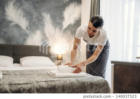Hotel housekeeping staff member arranging white towels on a neatly made bed in a modern guest room with decorative feather wall art and natural light Hotel housekeeping staff member arranging white towels on a neatly made bed in a modern guest room with decorative feather wall art and natural light 140037323