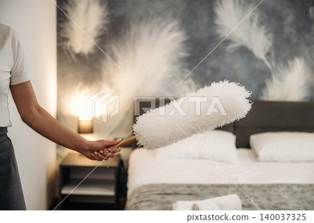 Hotel housekeeping staff member holding a feather duster while preparing a guest room with a neatly made bed and decorative wall art in the background Hotel housekeeping staff member holding a feather duster while preparing a guest room with a neatly made bed and decorative wall art in the background 140037325