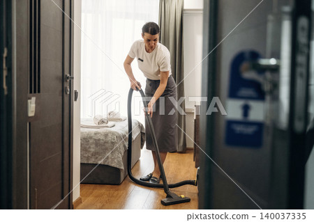 Hotel housekeeping staff member vacuuming carpeted floor in guest room, with neatly arranged towels on bed and natural light streaming through window Hotel housekeeping staff member vacuuming carpeted floor in guest room, with neatly arranged towels on bed and natural light streaming through window 140037335