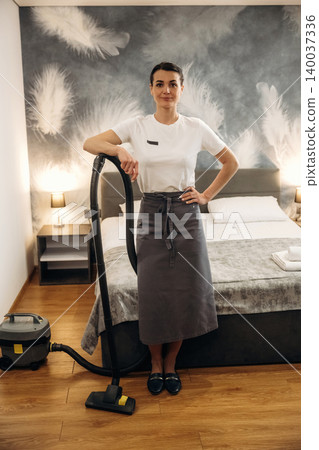 Hotel housekeeping staff member in gray apron stands with vacuum cleaner beside a neatly made bed in a modern hotel room with decorative wall design Hotel housekeeping staff member in gray apron stands with vacuum cleaner beside a neatly made bed in a modern hotel room with decorative wall design 140037336