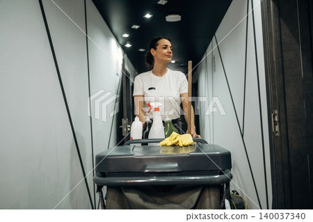 Female hotel staff member in a white shirt pushes a cleaning cart down a modern corridor with sleek walls and overhead lighting in a contemporary hotel setting Female hotel staff member in a white shirt pushes a cleaning cart down a modern corridor with sleek walls and overhead lighting in a contemporary hotel setting 140037340