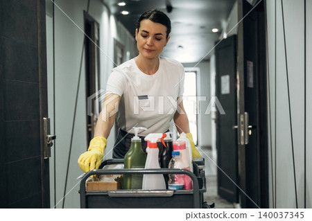 Female hotel housekeeper in white shirt and yellow gloves pushes cleaning cart filled with supplies down a modern hotel corridor with doors and lighting visible Female hotel housekeeper in white shirt and yellow gloves pushes cleaning cart filled with supplies down a modern hotel corridor with doors and lighting visible 140037345