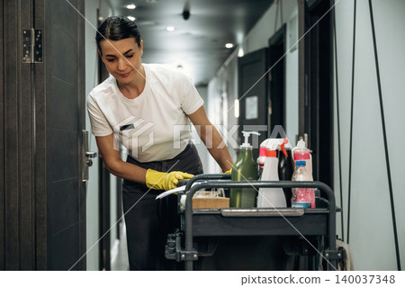 Hotel housekeeping staff member in uniform uses cleaning supplies on a cart while tidying up a corridor in a modern hotel environment Hotel housekeeping staff member in uniform uses cleaning supplies on a cart while tidying up a corridor in a modern hotel environment 140037348