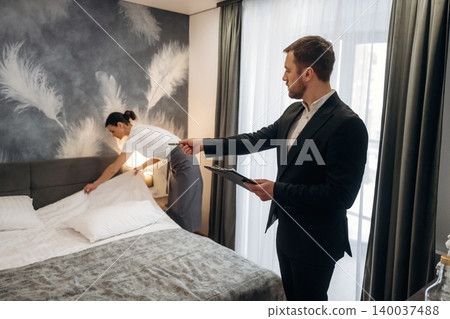 Hotel manager in formal attire observes housekeeping staff making bed in modern hotel room with elegant decor and large windows showcasing natural light Hotel manager in formal attire observes housekeeping staff making bed in modern hotel room with elegant decor and large windows showcasing natural light 140037488