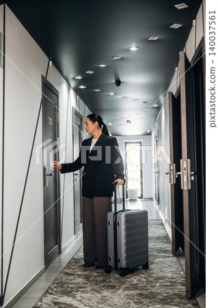 Female traveler in black blazer holding suitcase approaches hotel room door in modern corridor with stylish lighting and contemporary decor 140037561