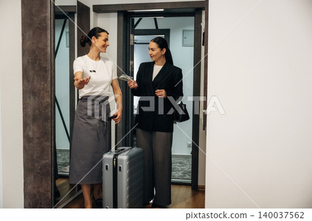 Hotel receptionist in gray skirt and white shirt greets a guest with a suitcase while standing in a modern lobby with mirrors and elevators in the background Hotel receptionist in gray skirt and white shirt greets a guest with a suitcase while standing in a modern lobby with mirrors and elevators in the background 140037562