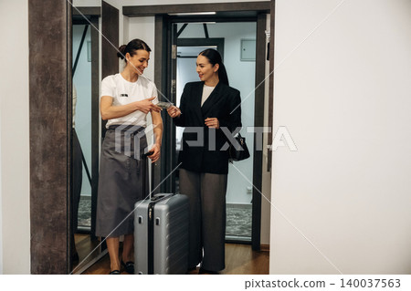 Hotel receptionist in gray uniform hands room key to a guest in a black blazer, standing by a suitcase near the elevator in a modern hotel lobby Hotel receptionist in gray uniform hands room key to a guest in a black blazer, standing by a suitcase near the elevator in a modern hotel lobby 140037563