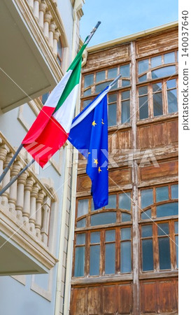Italian and EU flags against historic buildings in Baku, Azerbaijan. Flag of Italy and European Union together. Flag day celebration, patriotism. Embassies and consulates of countries Italian and EU flags against historic buildings in Baku, Azerbaijan. Flag of Italy and European Union together. Flag day celebration, patriotism. Embassies and consulates of countries 140037640