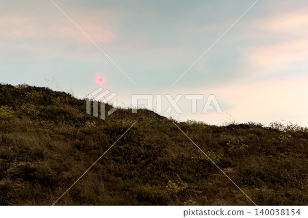 A natural landscape of grassy dunes under a subtle pastel sky with a distinct red sun A natural landscape of grassy dunes under a subtle pastel sky with a distinct red sun 140038154