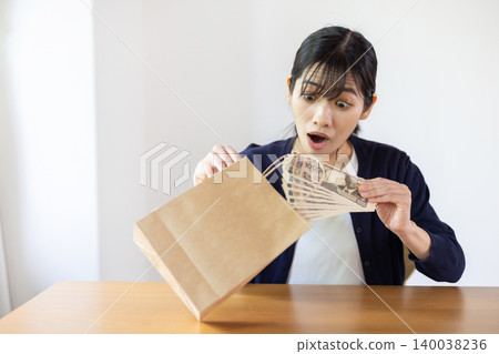 A young Japanese woman looks surprised as she takes a 10,000 yen bill out of a paper bag in a brightly lit room. 140038236