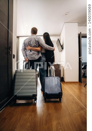 Couple standing together with luggage in a hotel room, embracing each other while facing the interior, wooden floor and modern furnishings visible in the background Couple standing together with luggage in a hotel room, embracing each other while facing the interior, wooden floor and modern furnishings visible in the background 140039069