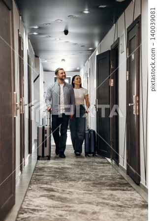 Couple walking down a hotel corridor with luggage, modern decor, and doors visible on both sides, showcasing a stylish and inviting atmosphere Couple walking down a hotel corridor with luggage, modern decor, and doors visible on both sides, showcasing a stylish and inviting atmosphere 140039081