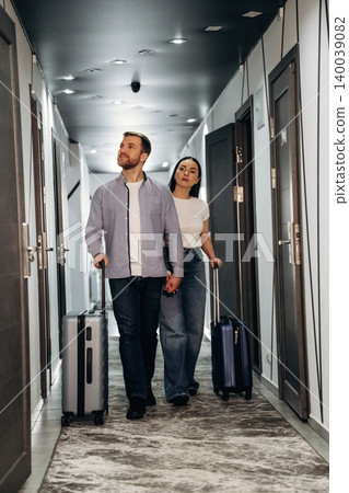 Couple walking hand in hand down a hotel corridor, each carrying a suitcase, with doors and modern lighting visible along the hallway Couple walking hand in hand down a hotel corridor, each carrying a suitcase, with doors and modern lighting visible along the hallway 140039082