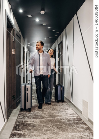 Couple walking down a hotel corridor with luggage, modern design elements visible, featuring gray walls, stylish lighting, and patterned carpet Couple walking down a hotel corridor with luggage, modern design elements visible, featuring gray walls, stylish lighting, and patterned carpet 140039084