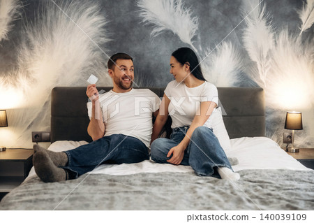 Young couple sitting on a hotel bed, smiling and engaging in conversation, with stylish feather-patterned wallpaper and bedside lamps in the background Young couple sitting on a hotel bed, smiling and engaging in conversation, with stylish feather-patterned wallpaper and bedside lamps in the background 140039109