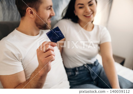 Couple sitting on a hotel bed, smiling and holding a blue condom package, wearing casual white t-shirts, with soft lighting and a cozy atmosphere in the background 140039112