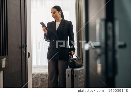 Businesswoman in black blazer checks smartphone while standing in hotel room with suitcase and luggage beside her, natural light streaming through window Businesswoman in black blazer checks smartphone while standing in hotel room with suitcase and luggage beside her, natural light streaming through window 140039148