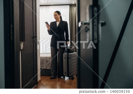Businesswoman in black suit holding smartphone and suitcase stands in hotel room doorway, modern decor and bed visible in the background Businesswoman in black suit holding smartphone and suitcase stands in hotel room doorway, modern decor and bed visible in the background 140039150