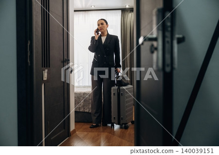 Businesswoman in a black suit stands in hotel room doorway holding a suitcase while talking on a mobile phone, with a bed and window visible in the background Businesswoman in a black suit stands in hotel room doorway holding a suitcase while talking on a mobile phone, with a bed and window visible in the background 140039151
