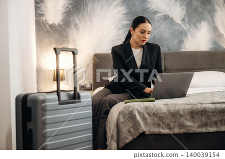 Businesswoman working on laptop in hotel room with suitcase beside bed, modern decor featuring feather wall art and soft lighting creating a professional atmosphere Businesswoman working on laptop in hotel room with suitcase beside bed, modern decor featuring feather wall art and soft lighting creating a professional atmosphere 140039154