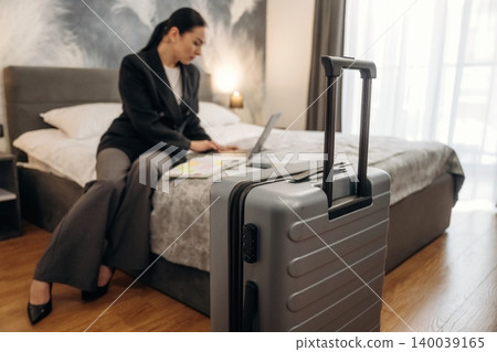 Businesswoman in black suit working on laptop while sitting on bed in hotel room with suitcase placed nearby, modern decor and natural light visible Businesswoman in black suit working on laptop while sitting on bed in hotel room with suitcase placed nearby, modern decor and natural light visible 140039165
