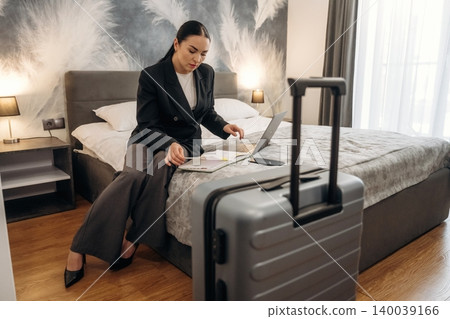 Businesswoman in black blazer sitting on hotel bed, reviewing travel documents and using laptop, with suitcase in foreground and modern decor in hotel room Businesswoman in black blazer sitting on hotel bed, reviewing travel documents and using laptop, with suitcase in foreground and modern decor in hotel room 140039166