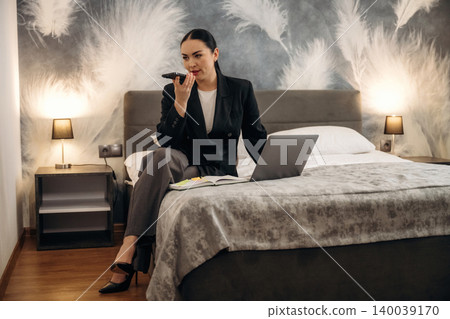 Businesswoman in black suit sitting on bed in hotel room, using smartphone while laptop and notebook are placed on bed with decorative wall in background Businesswoman in black suit sitting on bed in hotel room, using smartphone while laptop and notebook are placed on bed with decorative wall in background 140039170
